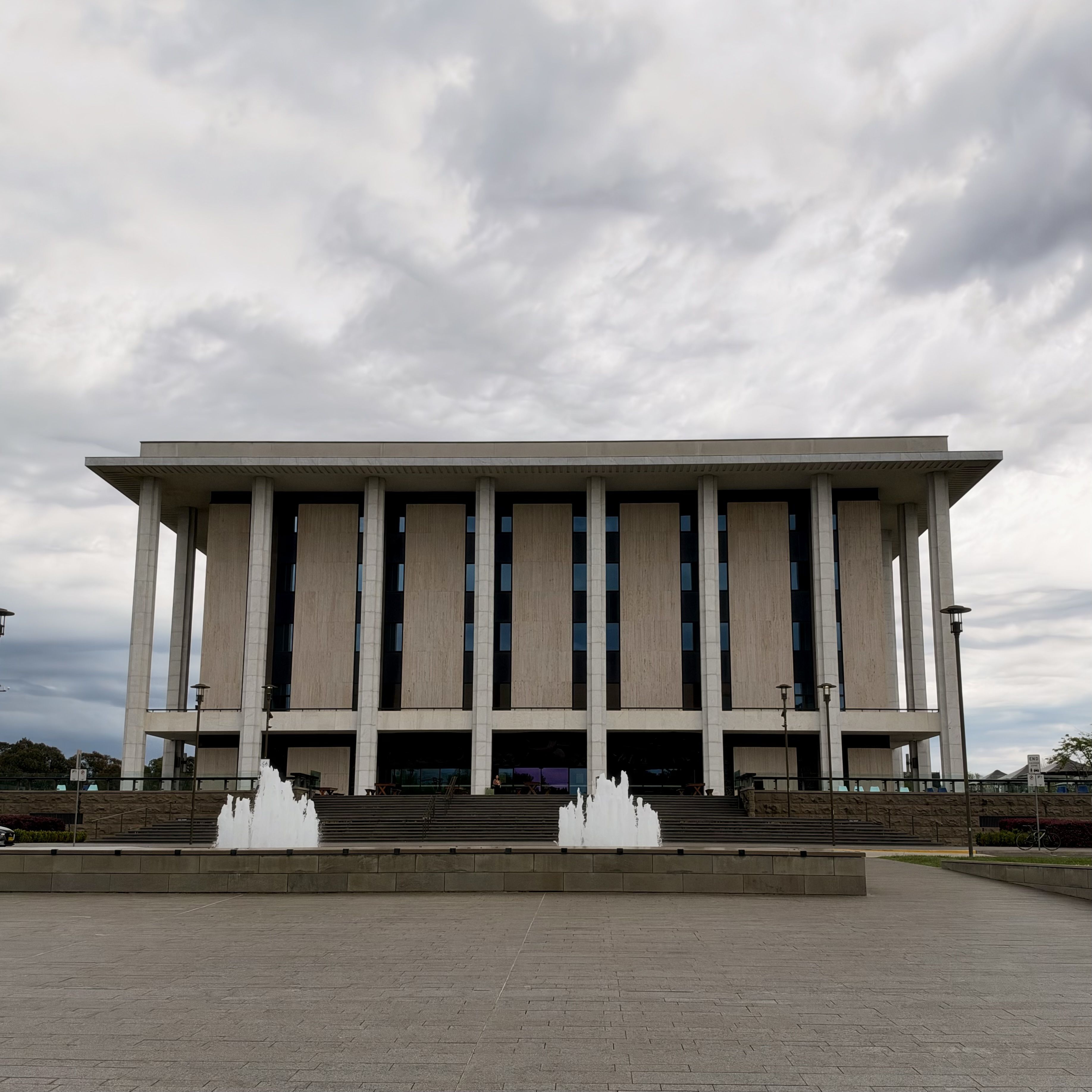 A large concrete building with tall columns and a fountain in front under a cloudy sky.