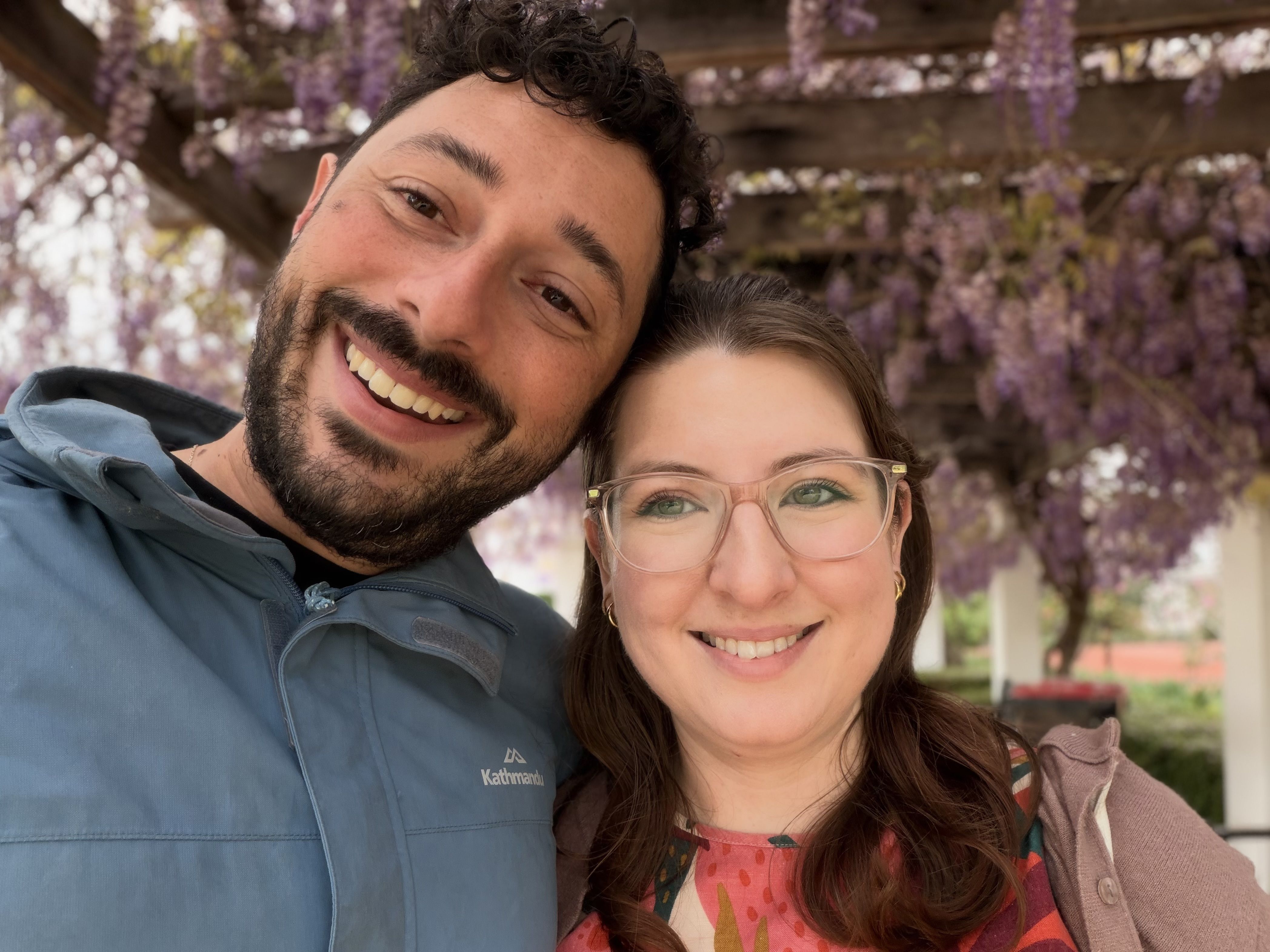 A man and a woman smiling together under a purple-flowered pergola.