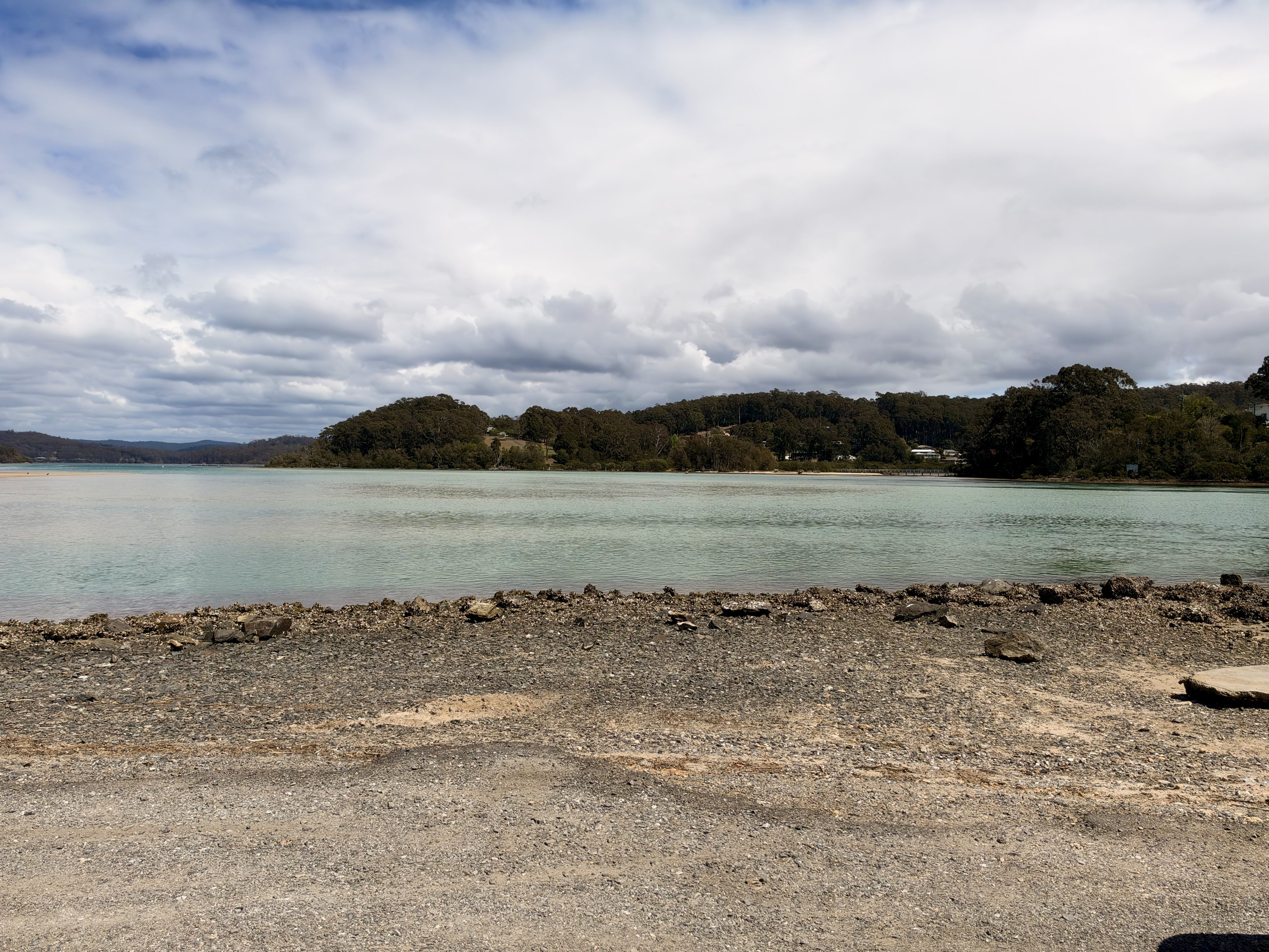 A rocky beach with a lake and trees under a cloudy sky.