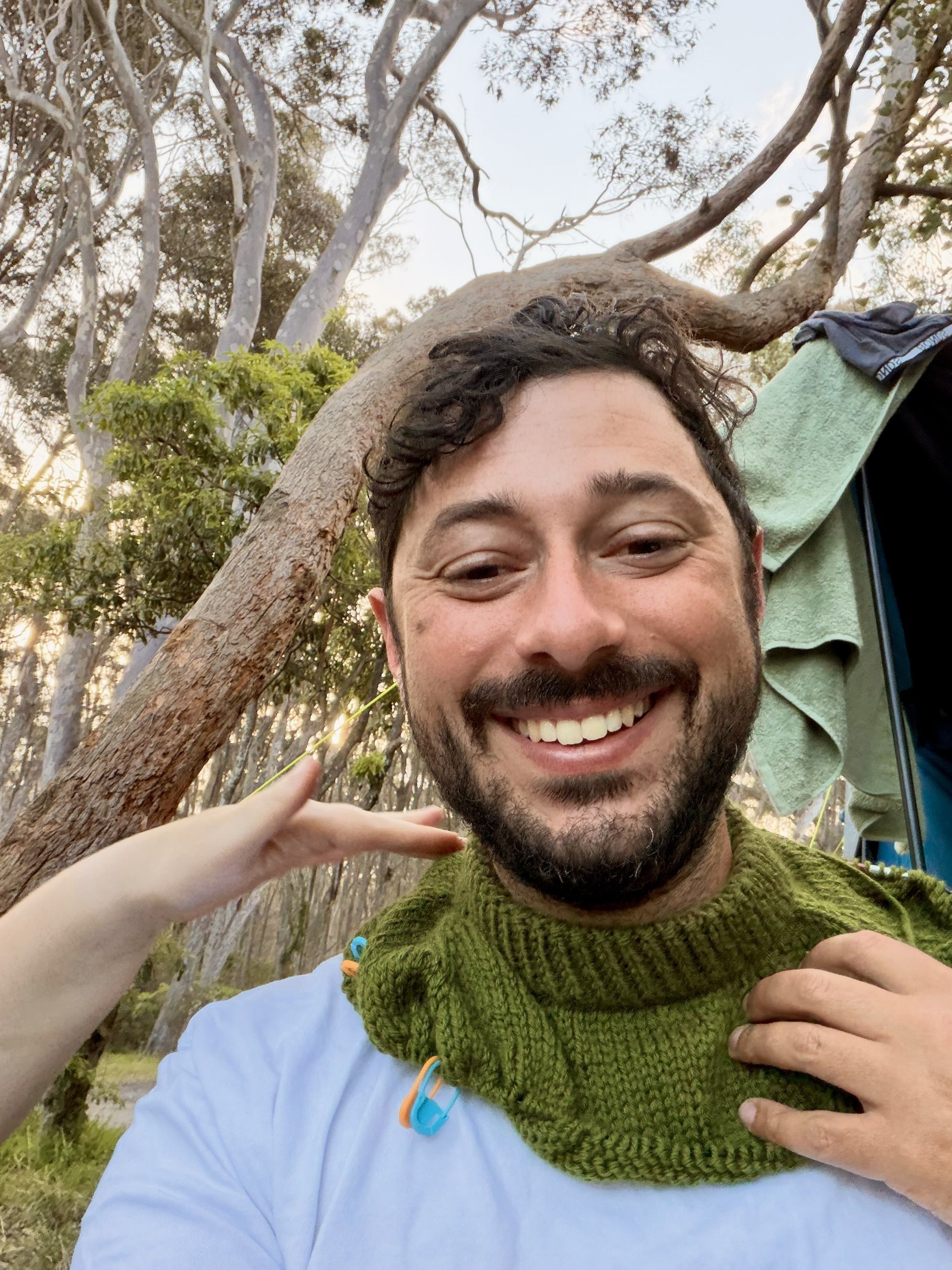 A man in a blue shirt and a green knitted scarf smiling in front of trees and a tent.