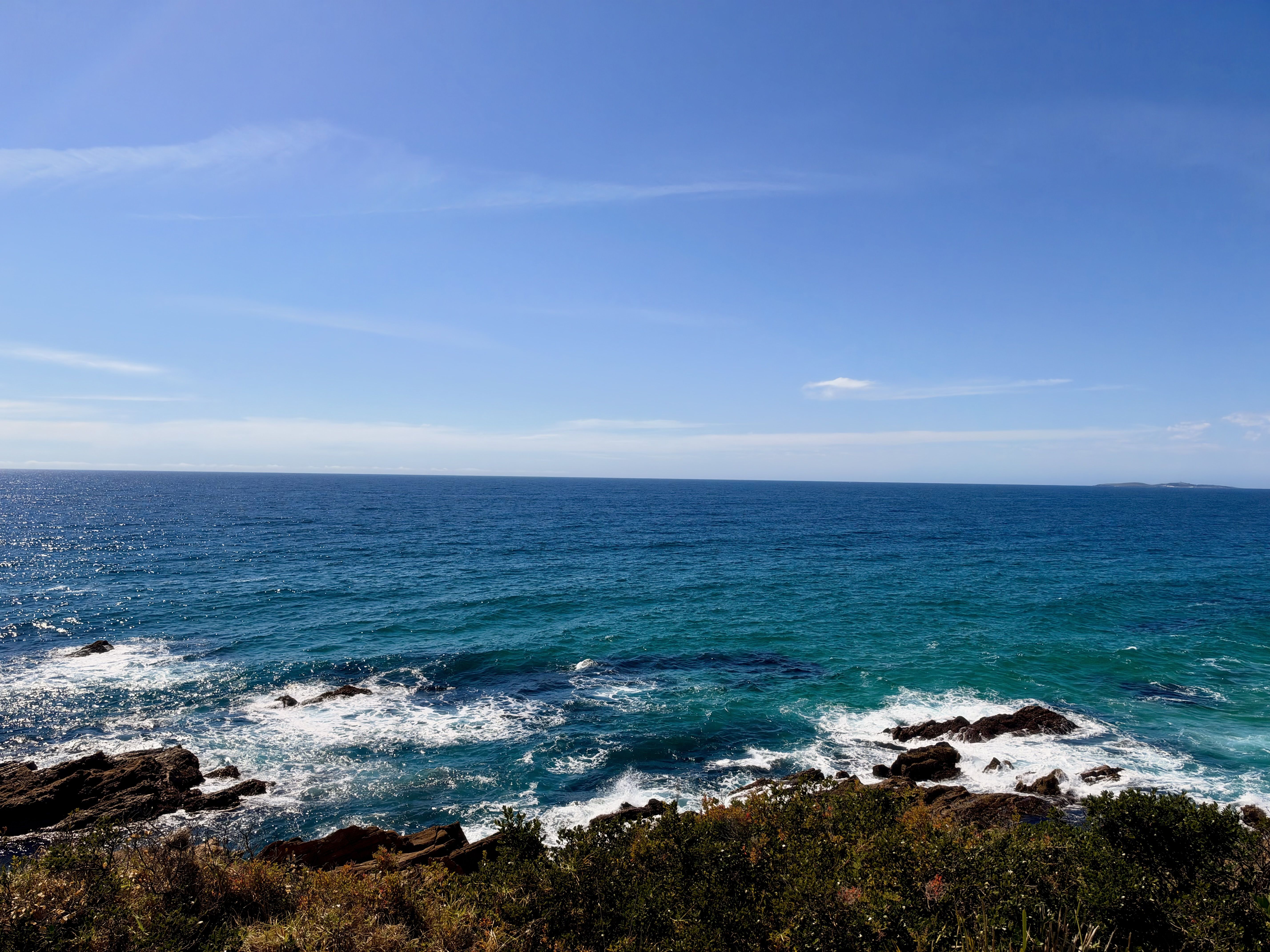 A beach with blue water, rocks, and greenery under a clear blue sky.