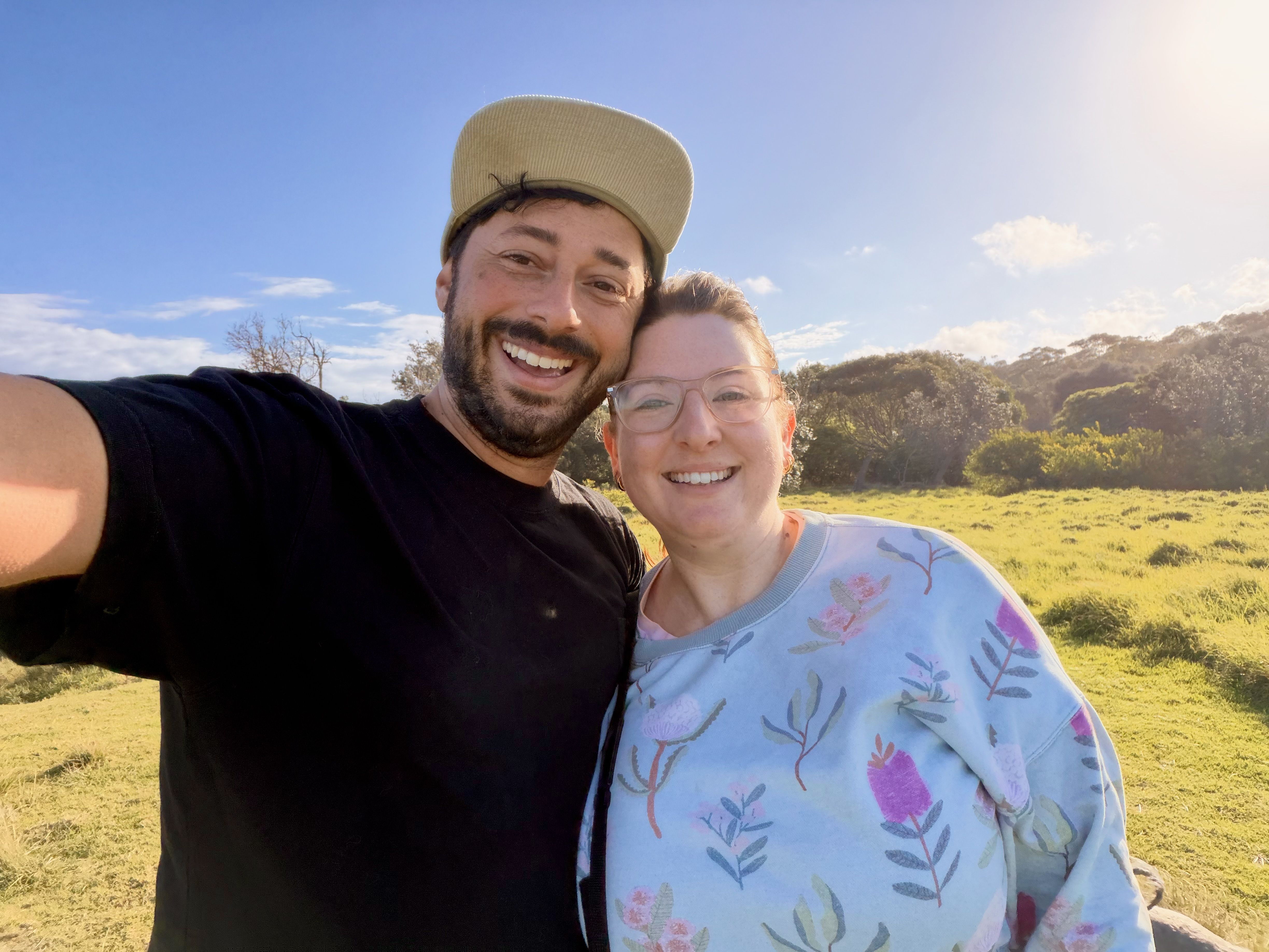 Two people smiling in a grassy field under a clear blue sky.