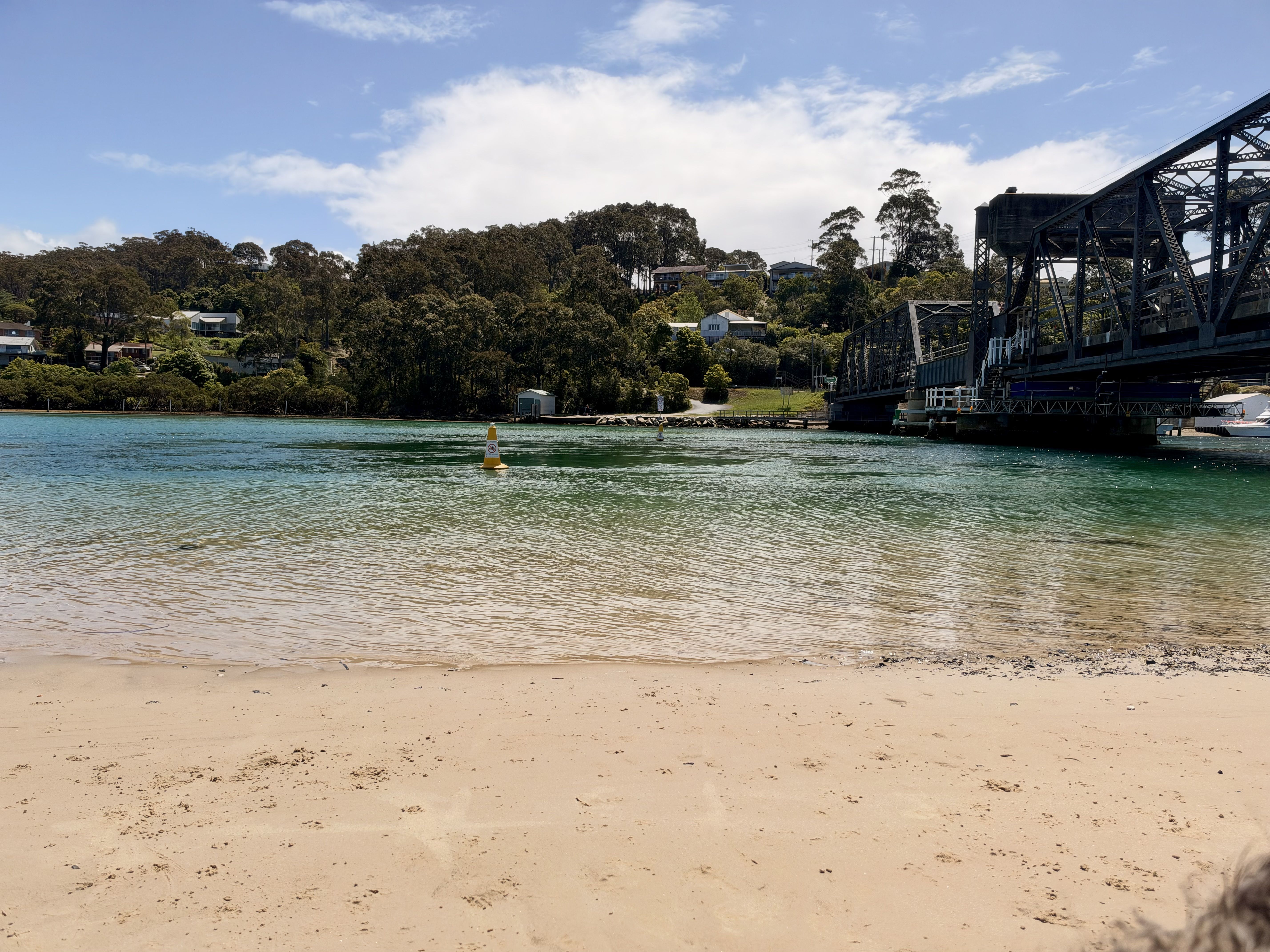 A sandy beach with a body of water, a bridge, and a hillside with trees and houses.