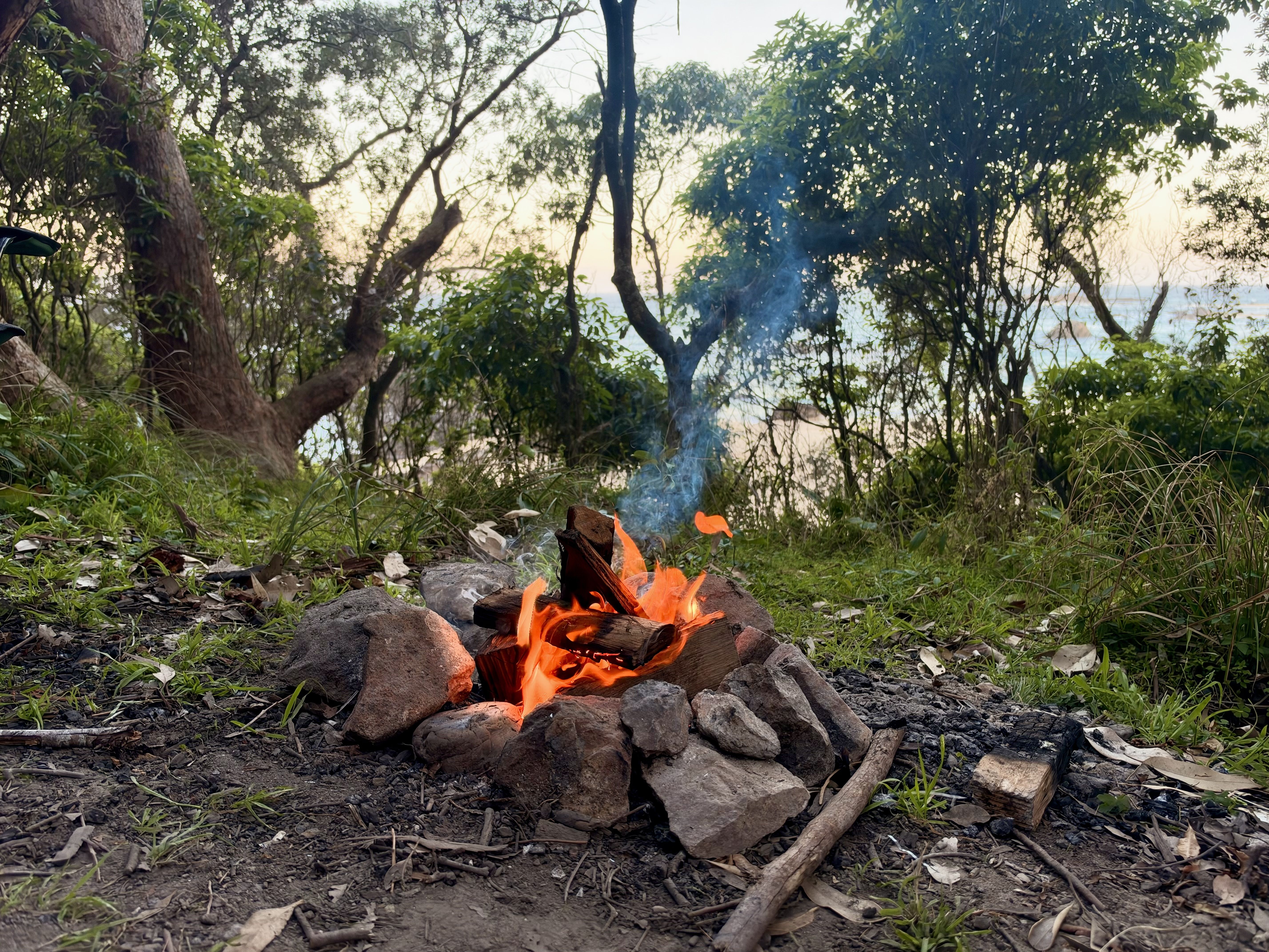 A campfire with flames and smoke surrounded by rocks and trees.