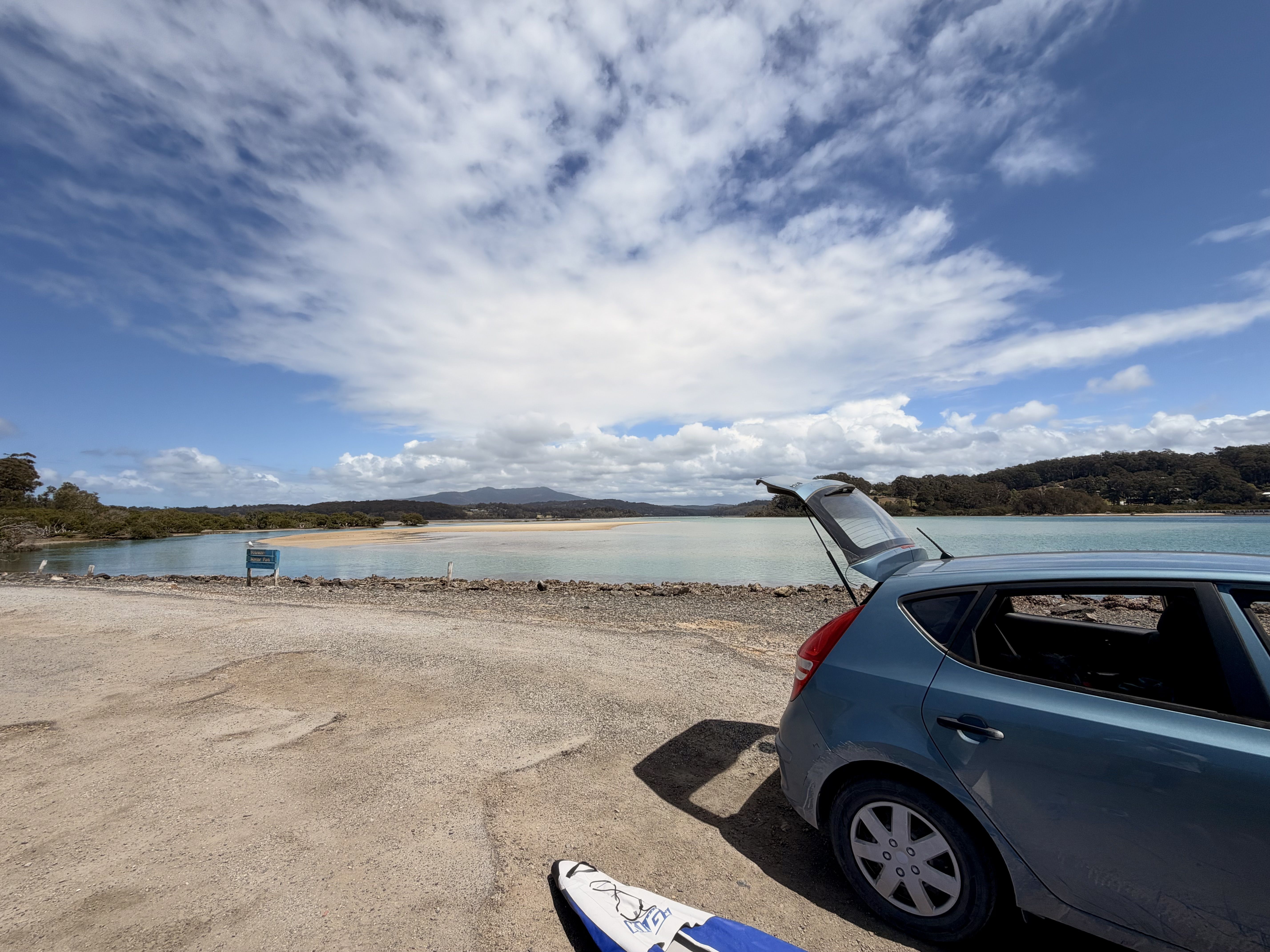 Blue car with open trunk on sandy area near beach with mountains under cloudy sky.