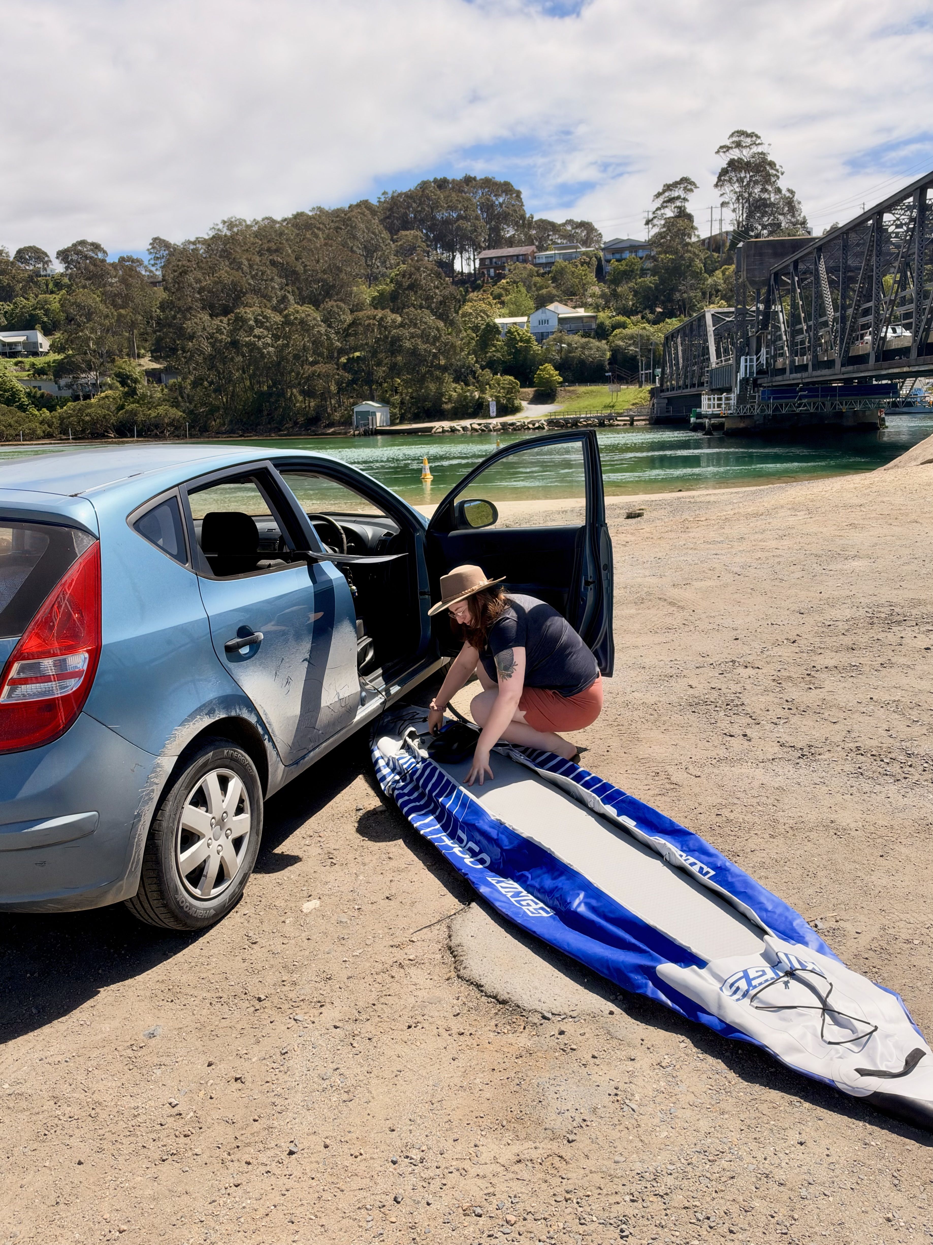 A woman in a hat leans into a blue car, holding a surfboard on a sandy area near water.