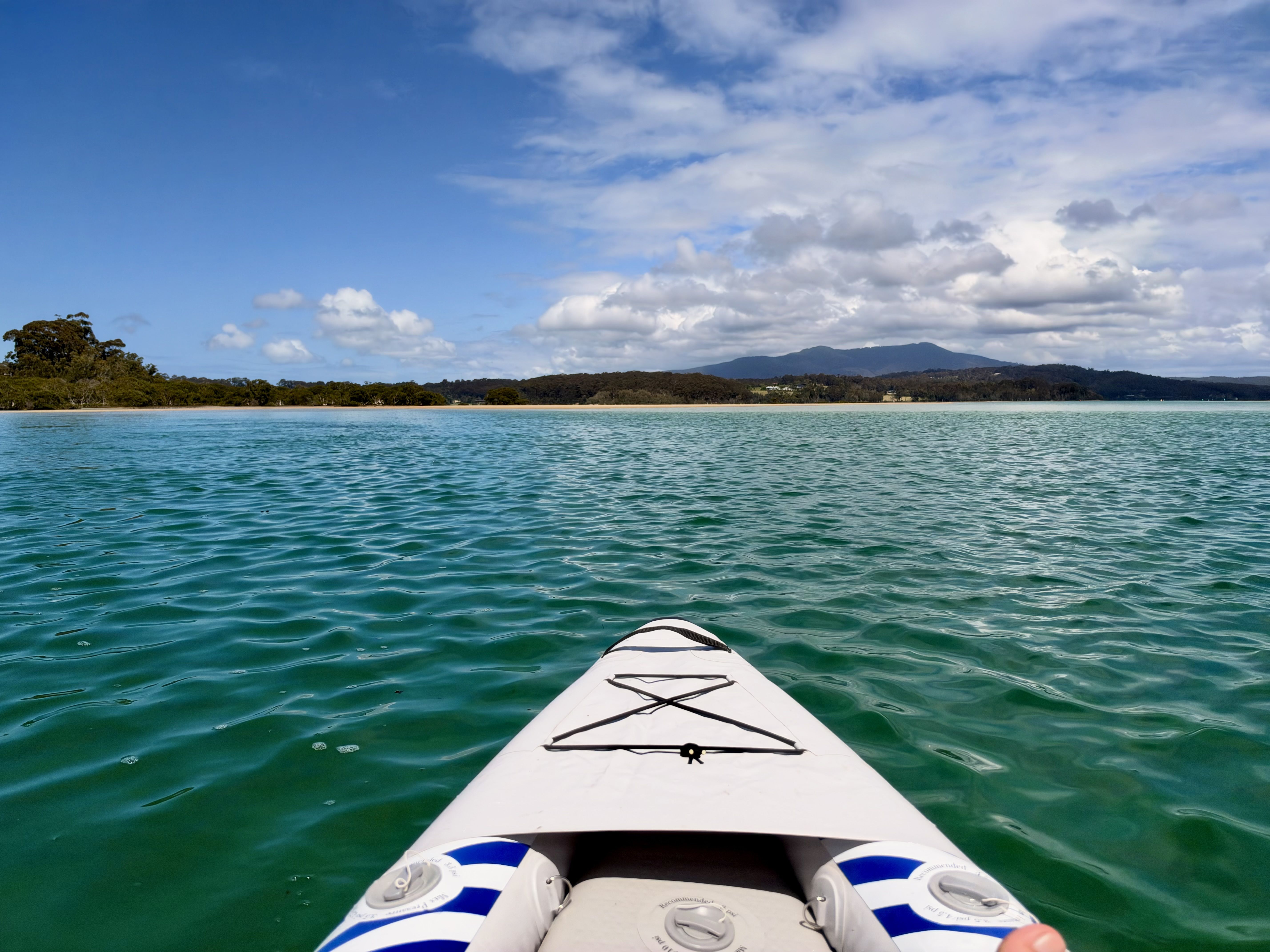 A white kayak with blue accents on turquoise water, with a distant shoreline and mountains under a cloudy blue sky.