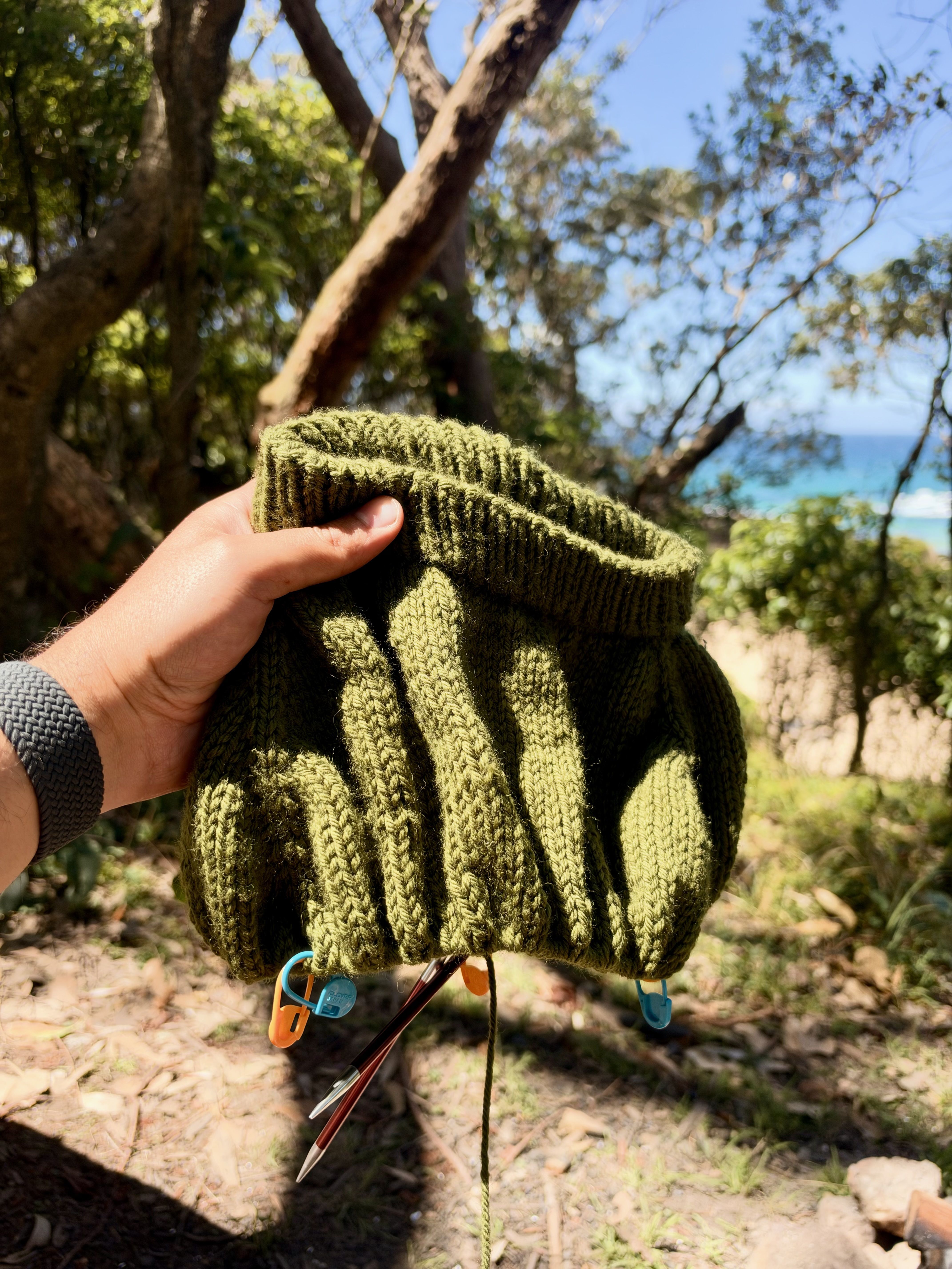 A person holds a green knitted object with colorful clips in a natural setting with trees and water.