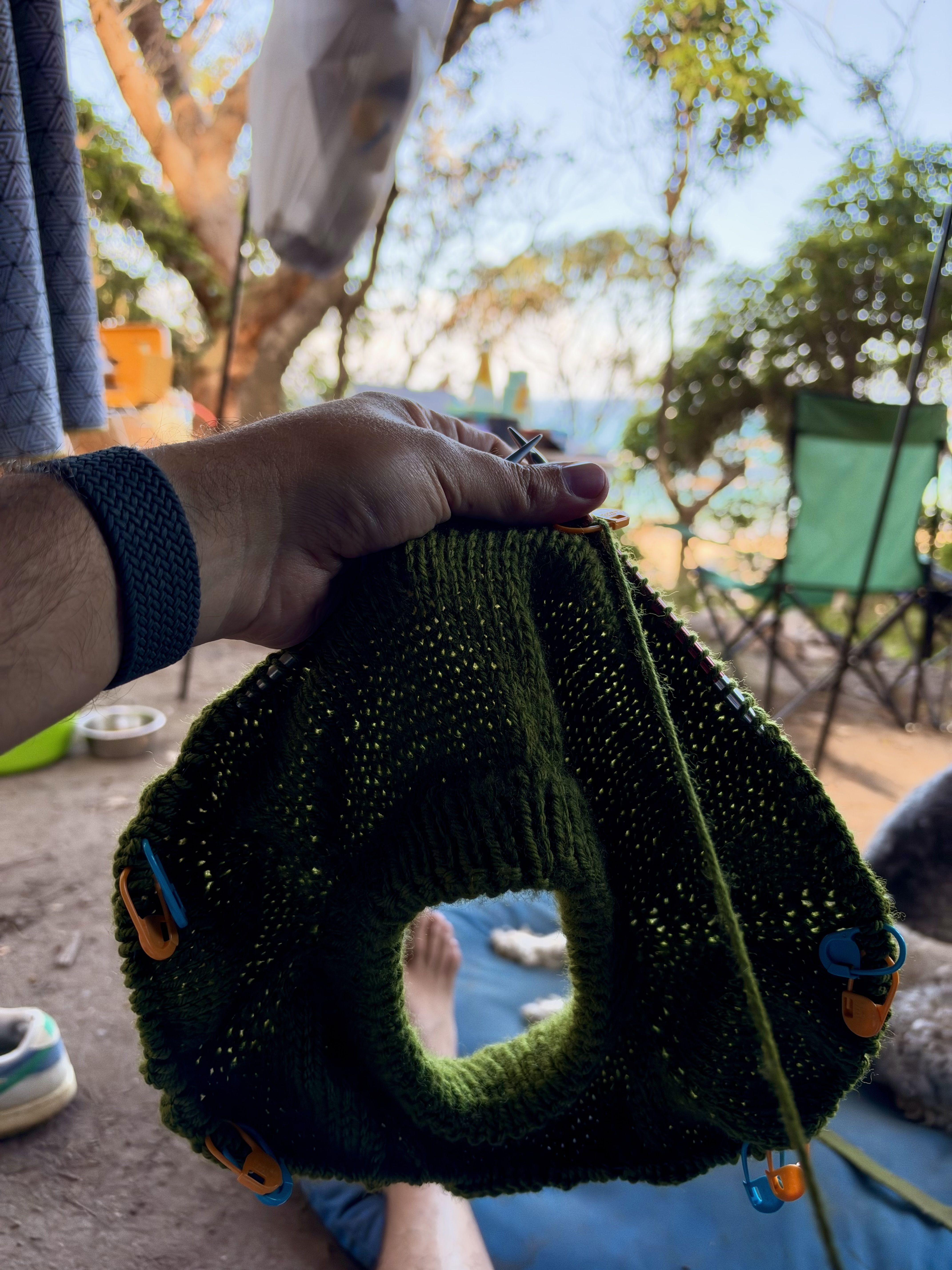 A person holds a green knitted object with colorful beads near a chair and trees.